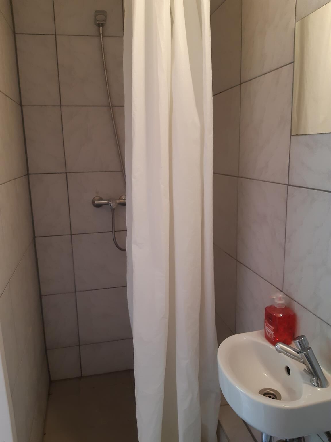 Casa de banho com chuveiro, lavatório e frasco de sabonete líquido vermelho.