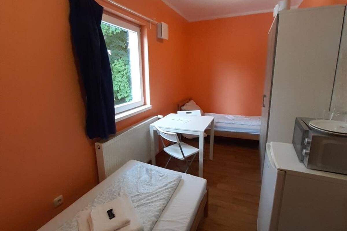 Een kleine kamer met een bed, een tafeltje, een magnetron en een raam met gordijnen.