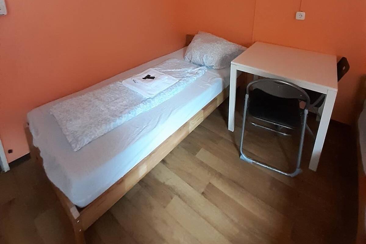 Een kleine eenpersoonskamer met een oranje muur, een houten vloer en een wit bureau met stoel.