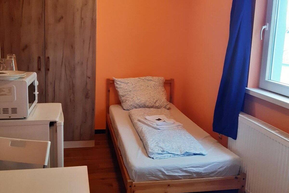 Une petite chambre avec un lit simple, une armoire en bois, une table avec un micro-ondes et une fenêtre avec un rideau bleu.