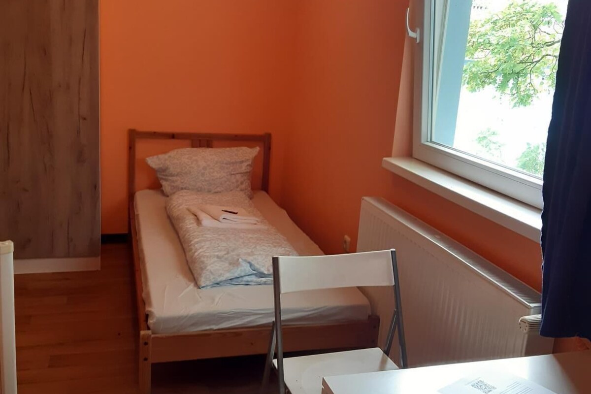 Une petite chambre avec un lit simple, un lit en bois, une chaise blanche, une table et une fenêtre donnant sur des arbres.