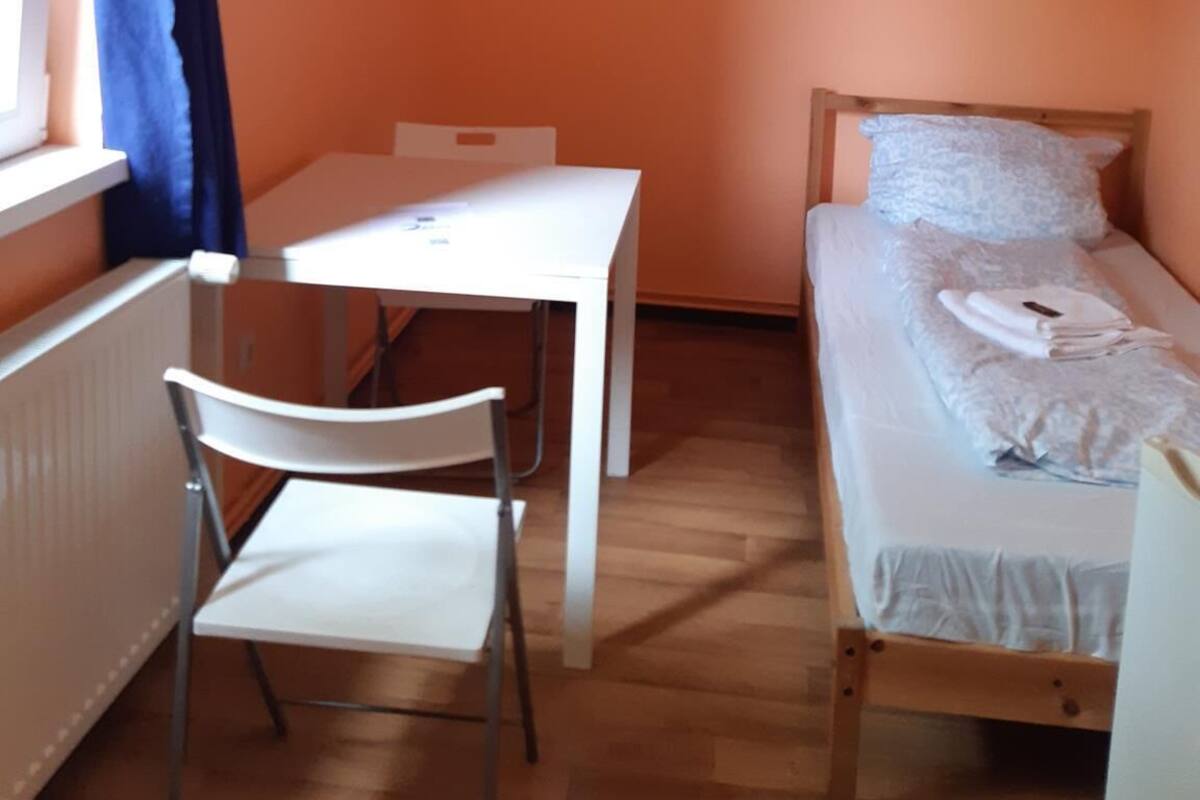 Una habitación pequeña con una pared naranja, un escritorio blanco con una silla y una cama individual con colchón y almohada blancos.