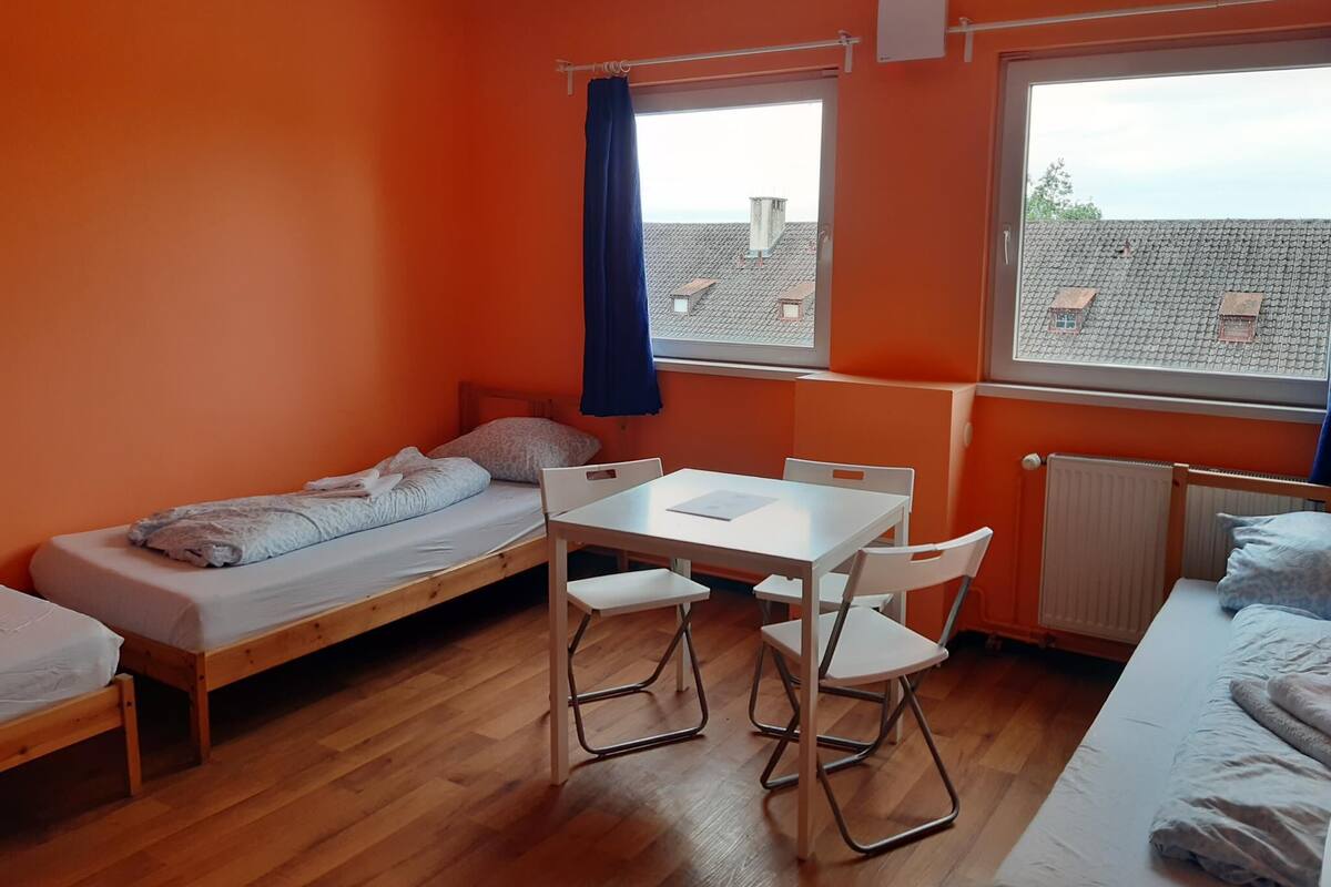 Un dormitorio con dos camas individuales, una mesita con sillas y una ventana con vistas a tejados.