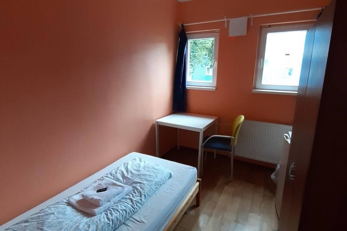 Een kleine kamer met een enkel bed, een houten vloer, een bureau, een stoel en een raam met gordijnen.