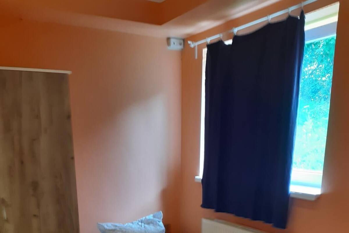 Een kleine kamer met een enkel bed, een houten vloer, een witte tafel en een raam met een donker gordijn.
