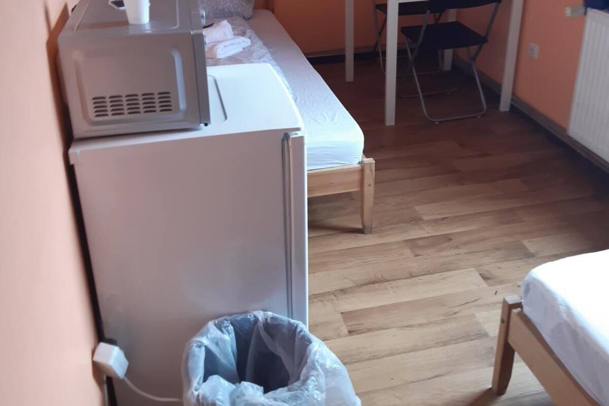 Una habitación pequeña con una cama, una mesa, una silla, un refrigerador y un cesto de basura.