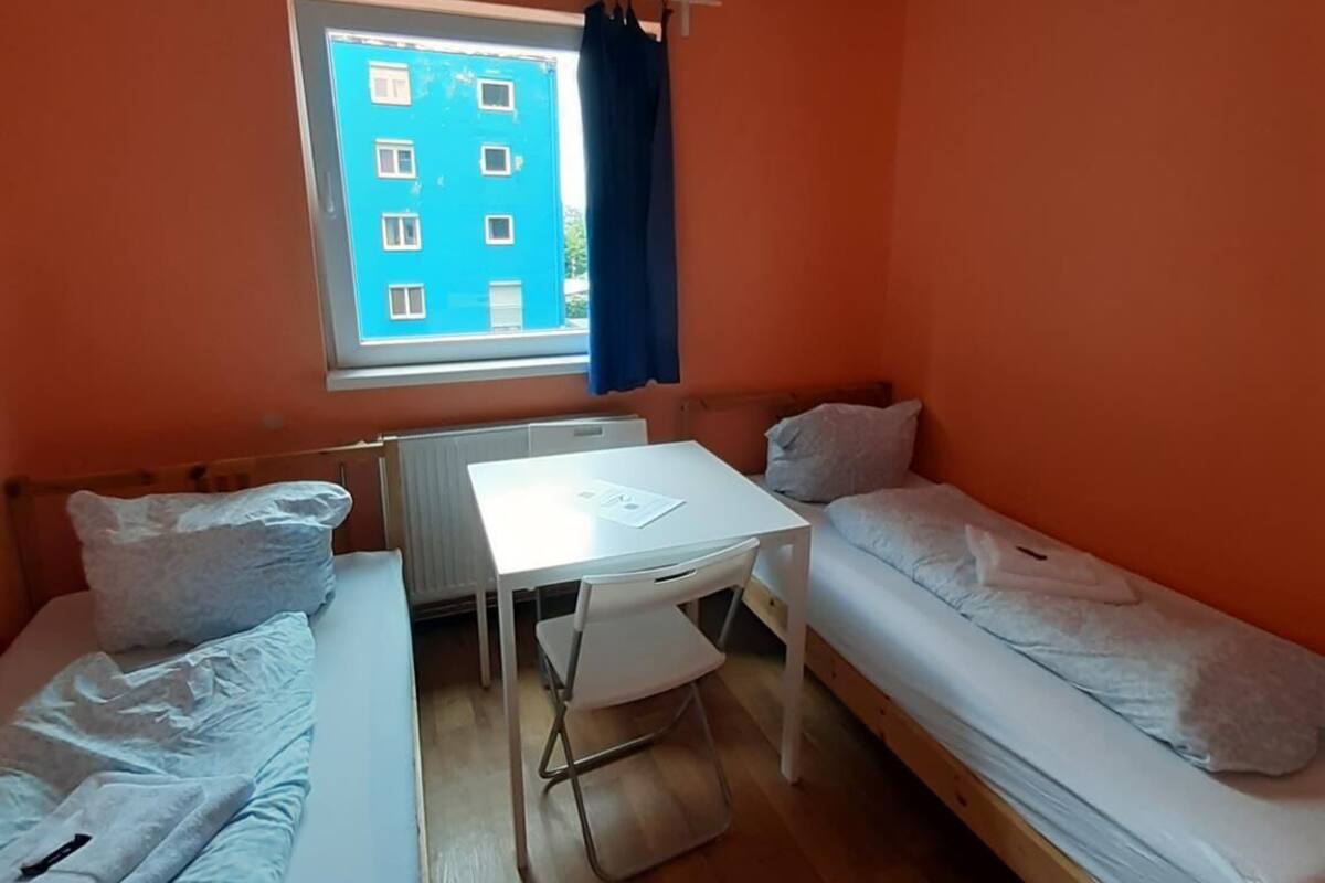 Een kleine kamer met oranje wanden, twee eenpersoonsbedden, een tafeltje en een raam met uitzicht op een blauw gebouw.