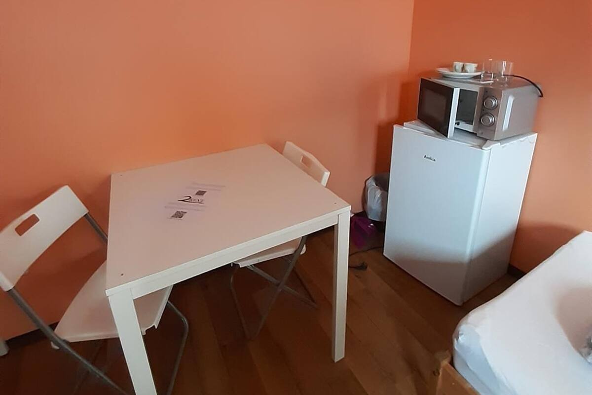 Een kleine kamer met een witte tafel, een witte stoel, een magnetron en een bed.