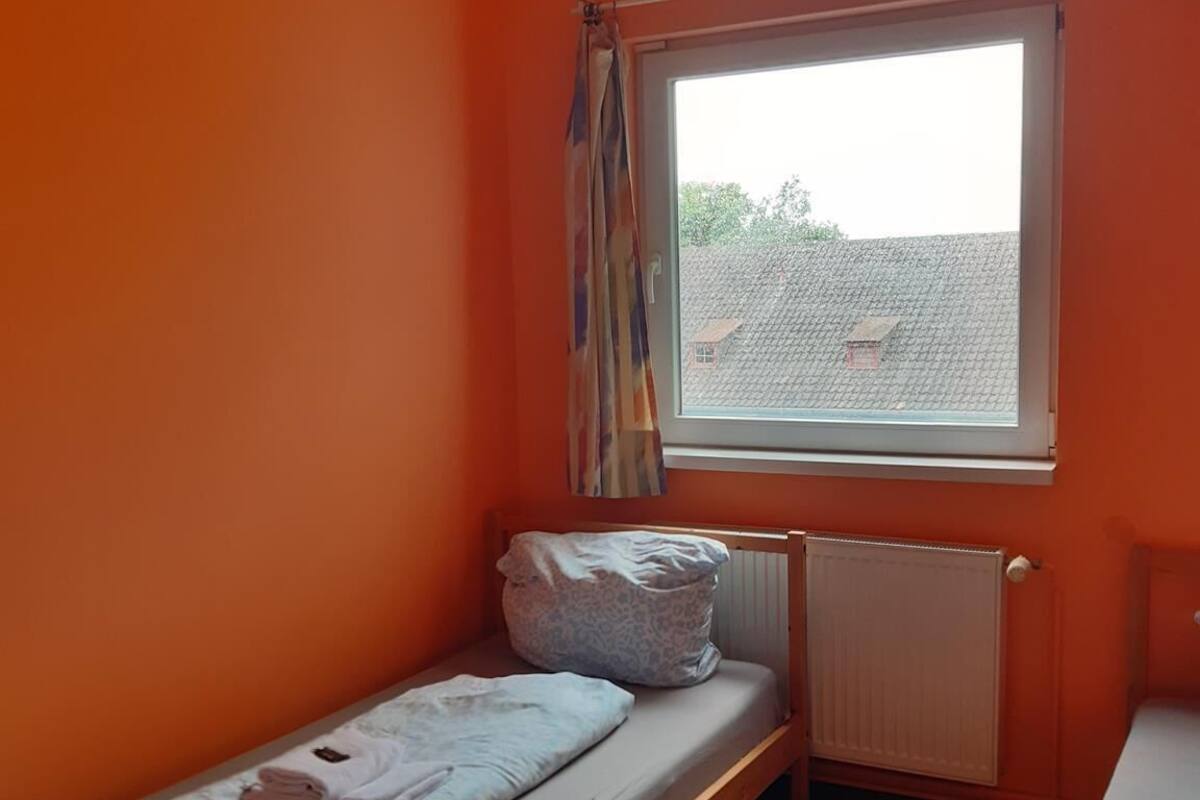 Una habitación pequeña con paredes anaranjadas, una única ventana, suelo de madera y una cama con un radiador debajo.