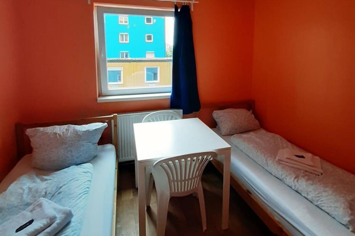 Een kleine, lichte kamer met oranje wanden, twee eenpersoonsbedden, een witte tafel en een stoel.
