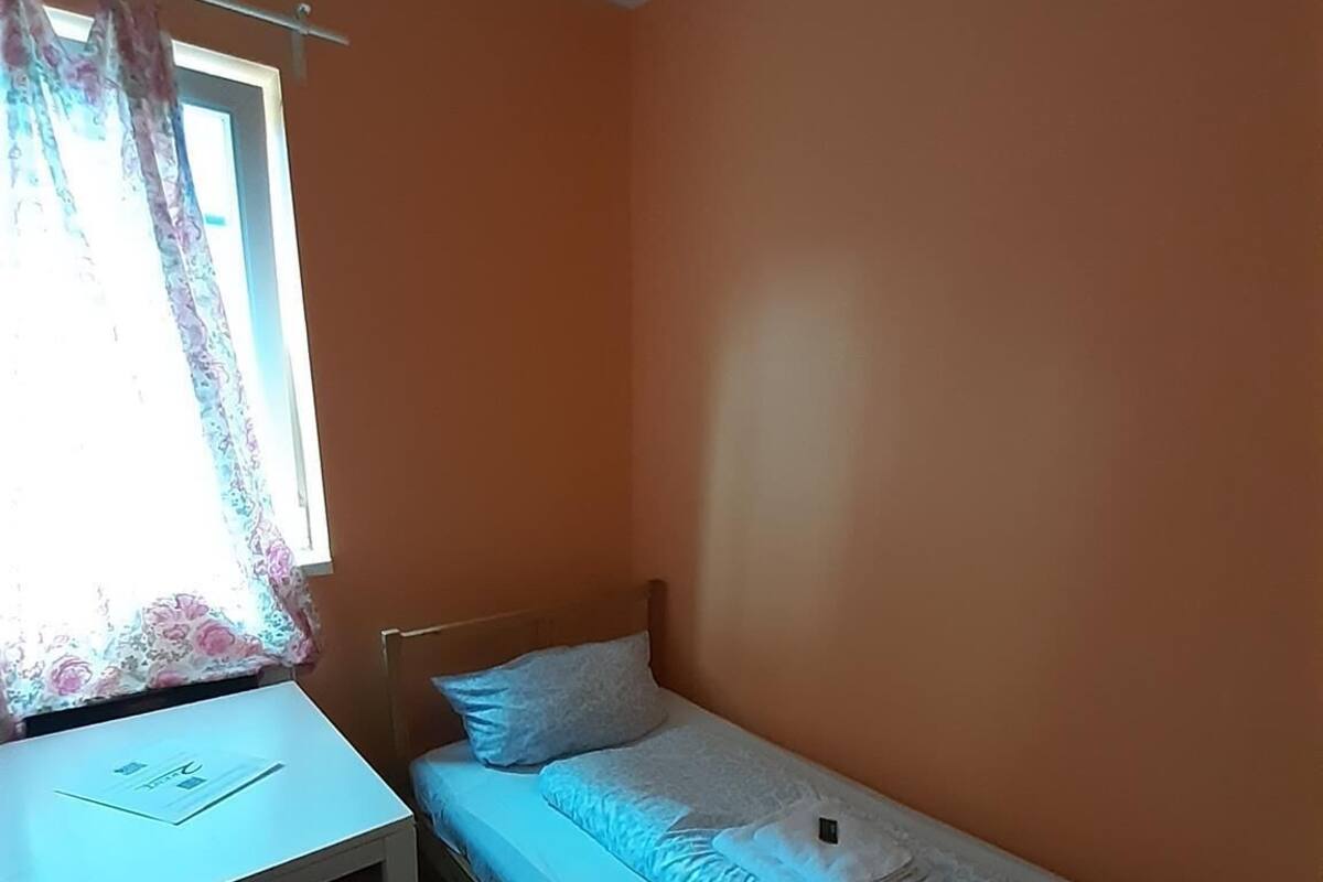 Een kleine eenpersoonskamer met een oranje muur, een bureau met stoel en een raam met bloemen gordijnen.