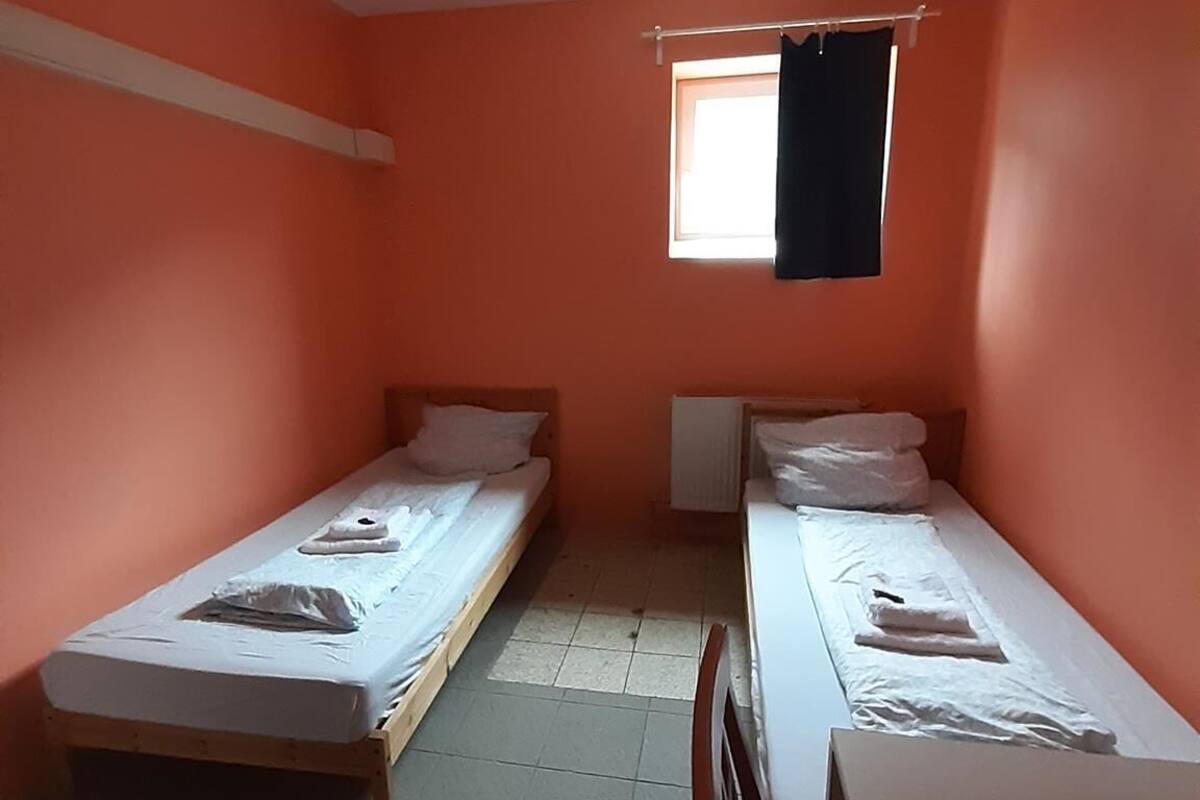 Een kleine, eenvoudige kamer met twee eenpersoonsbedden, een raam met gordijn en een radiator.