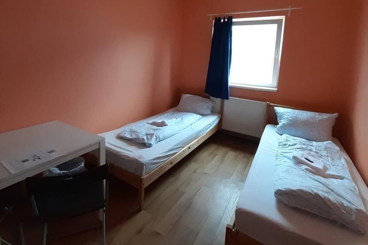 Een kleine kamer met oranje wanden, twee enkele bedden, een bureau en een raam met blauwe gordijnen.