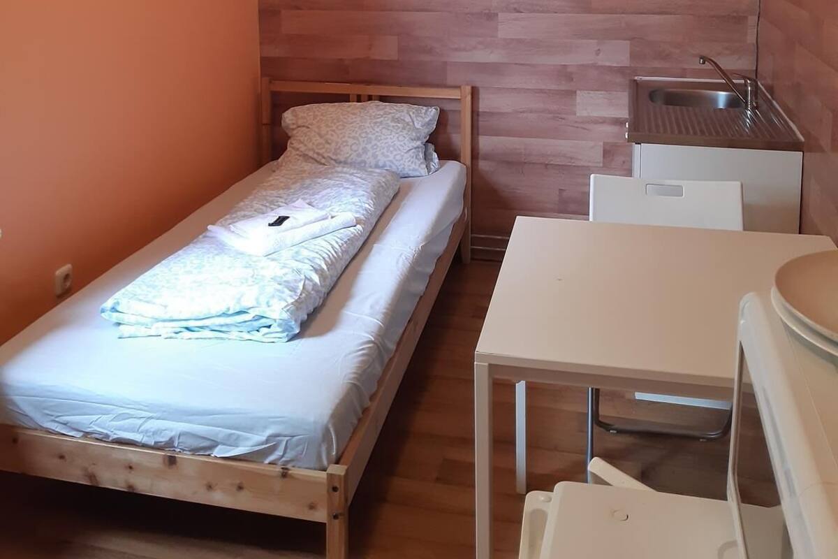 Una habitación pequeña con cama individual, cabecera de madera, una kitchenette compacta con fregadero y una mesa blanca.