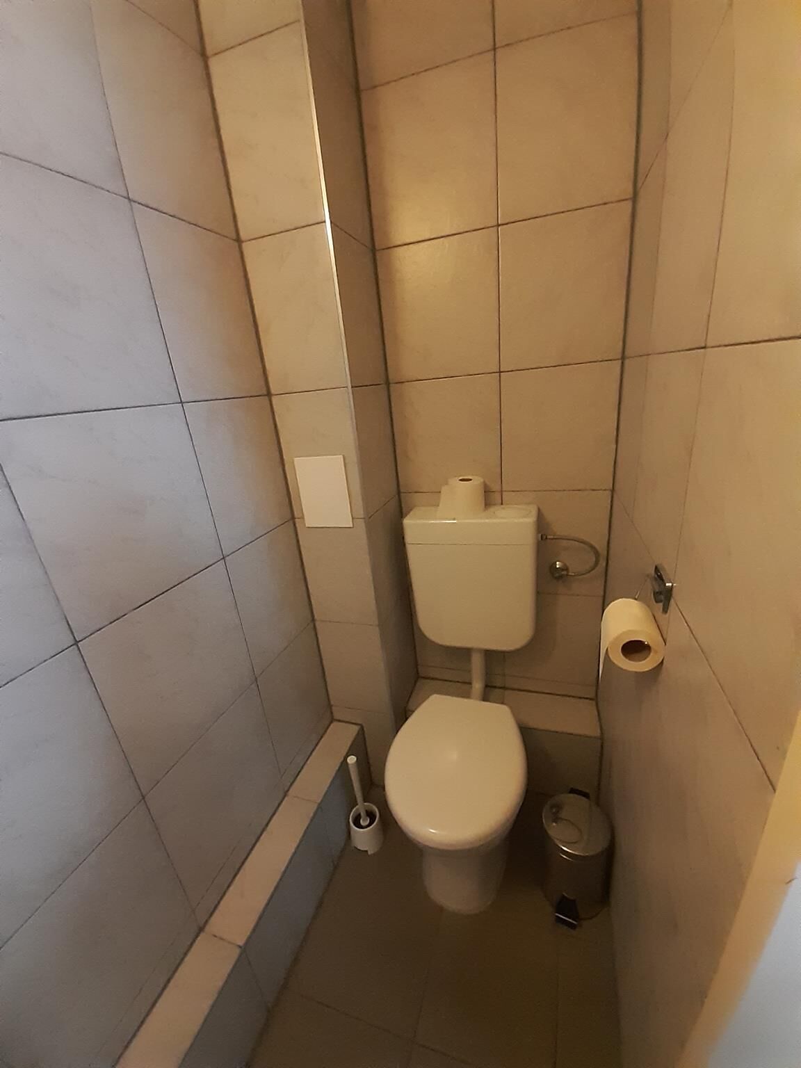 A small, tiled bathroom with a toilet, a trash can, and a toilet paper holder.