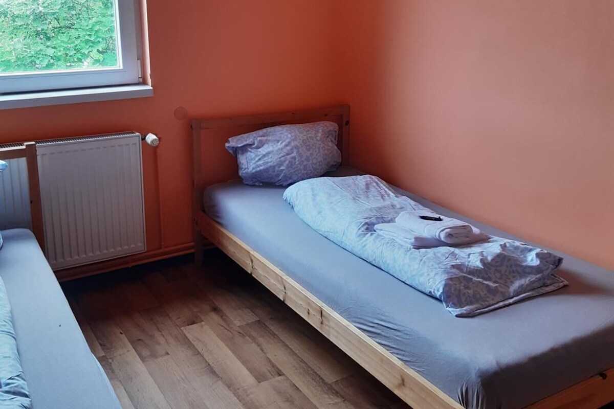 Une petite chambre avec un lit simple, un plancher en bois, des murs orange, une fenêtre donnant sur la verdure et un radiateur.
