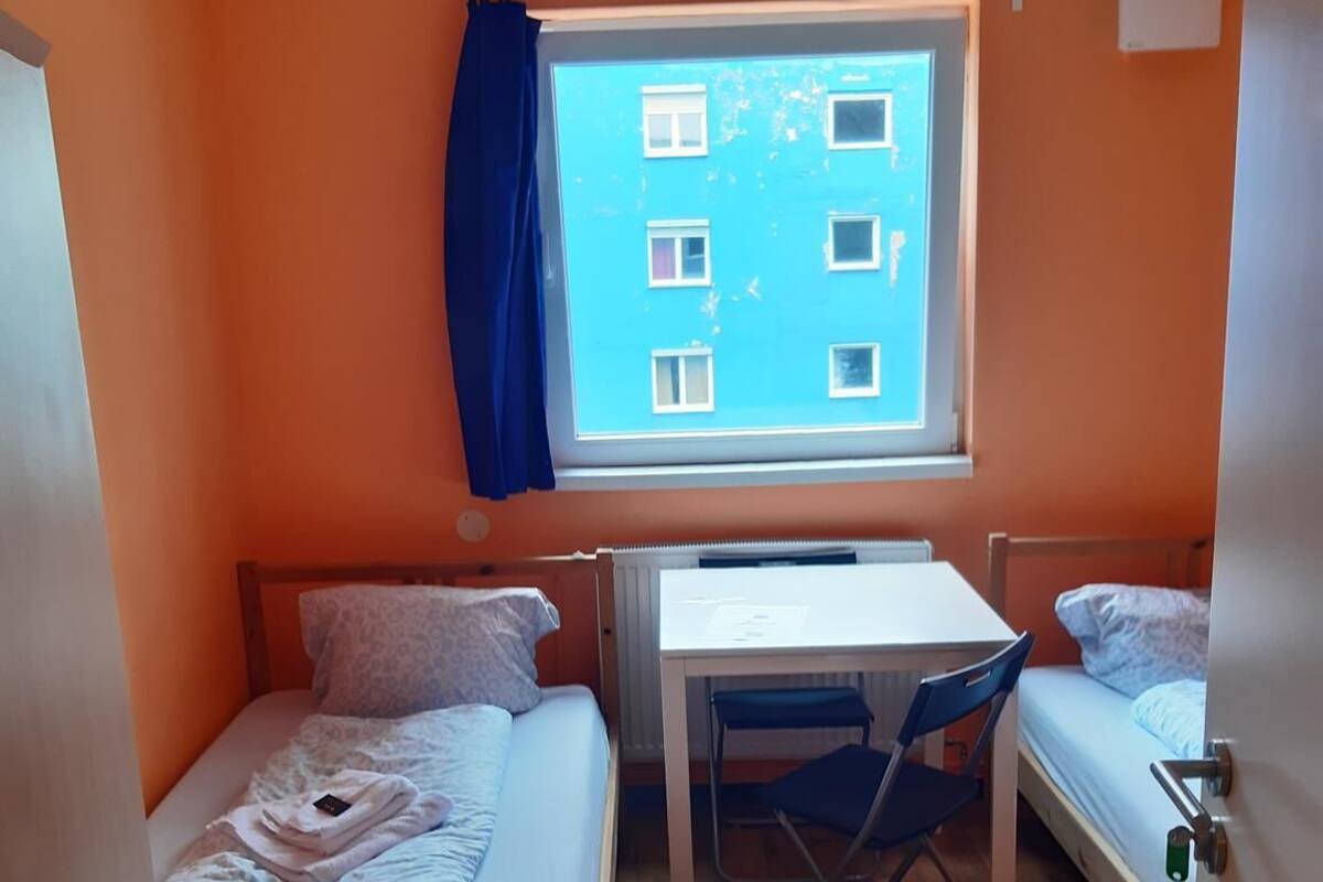 Een kleine kamer met oranje wanden, twee bedden, een bureau en een raam met uitzicht op een gebouw.