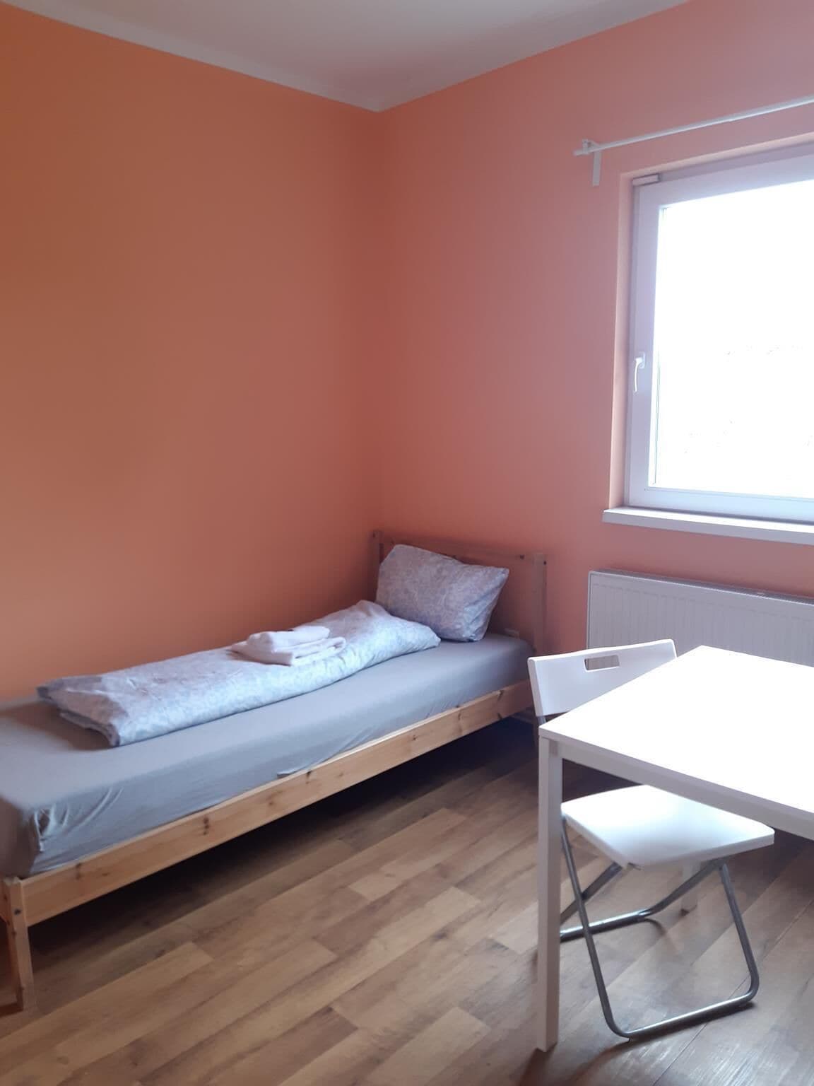 Een kleine kamer met een eenpersoonsbed, een witte tafel en een raam.