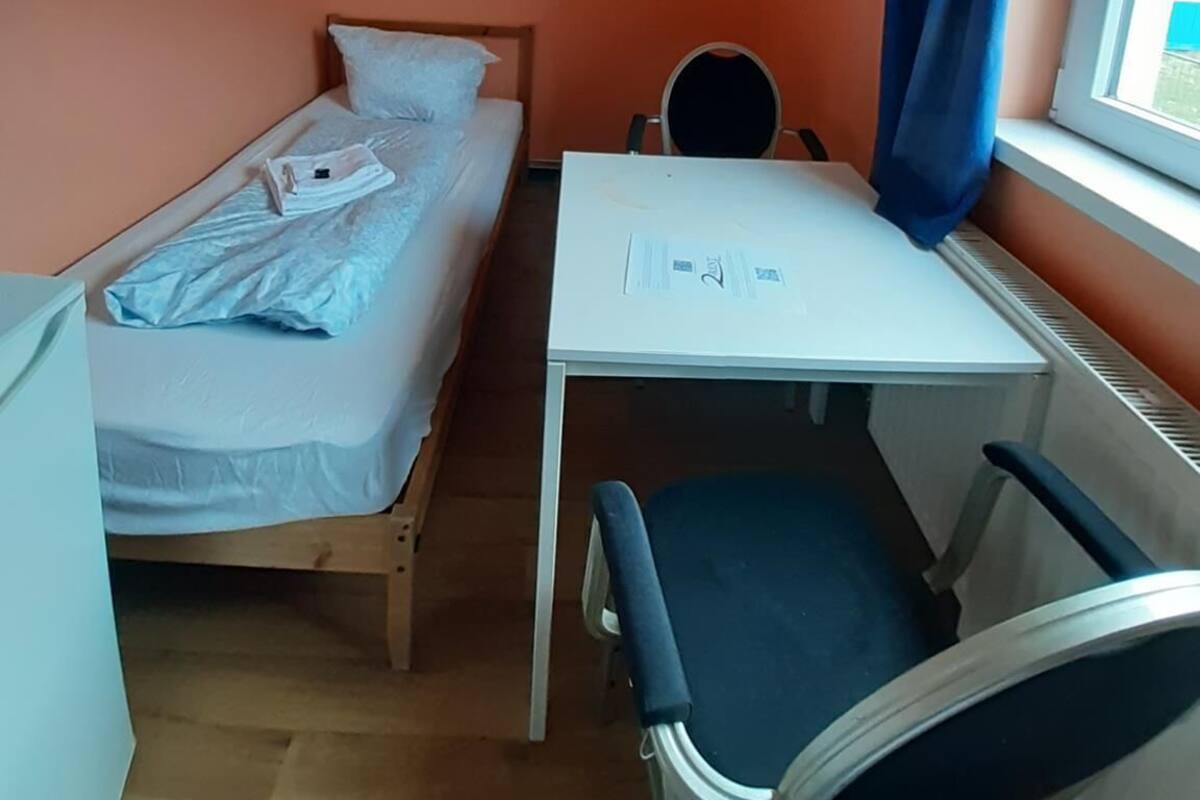 Een kleine kamer met een enkel bed, een bureau en stoel, een raam met blauwe gordijnen en een plafondlamp.