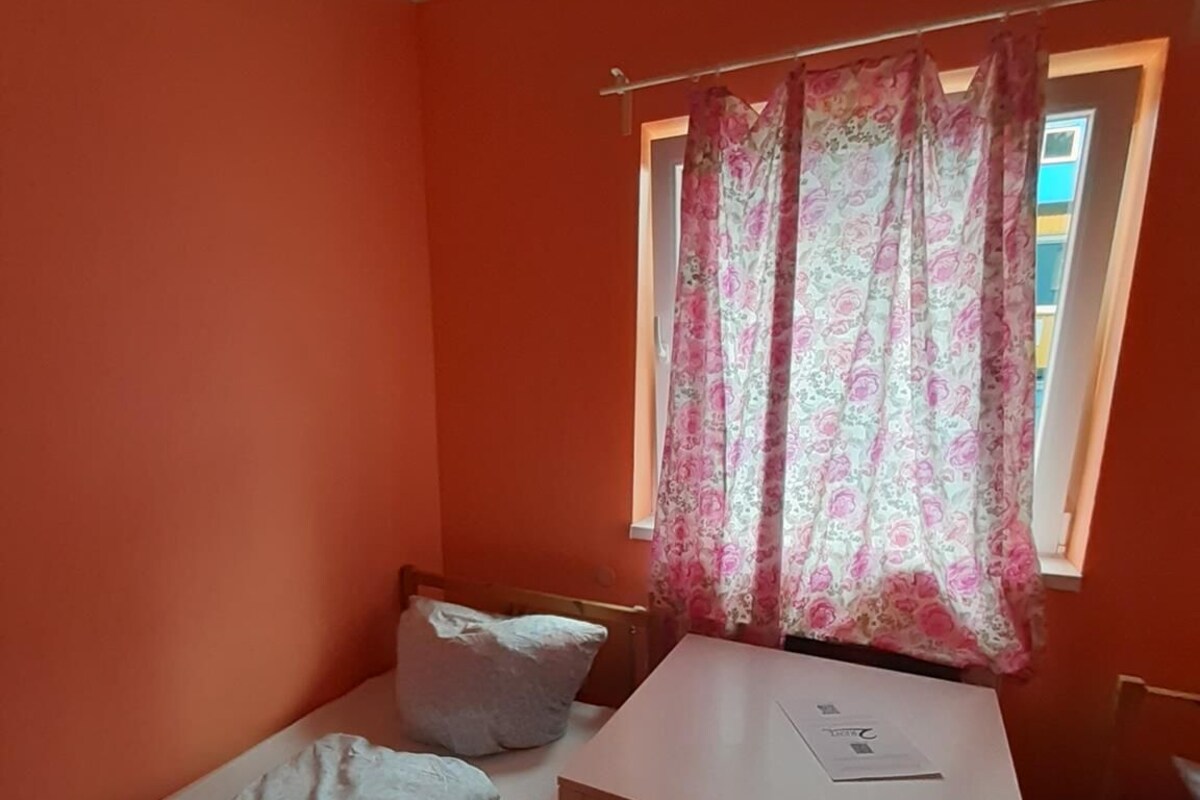 Une petite pièce avec un lit simple, une petite table et une fenêtre ornée de rideaux à motifs floraux.