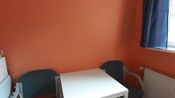 Een kamer met een oranje muur, een witte tafel, twee blauwe stoelen en een raam met blauwe gordijnen.