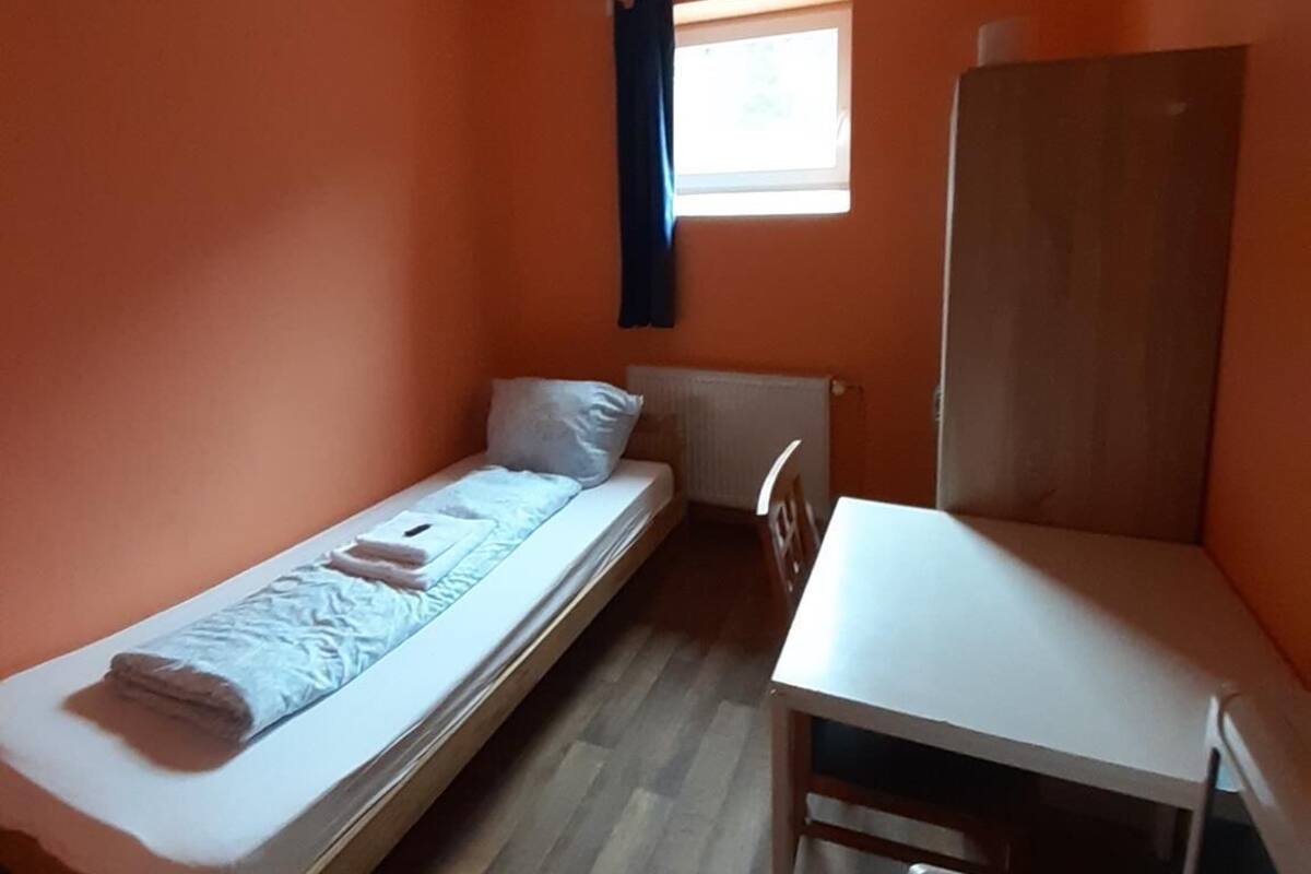 Een kleine eenpersoonskamer met een oranje wand, een raam met blauwe gordijnen, een wit bureau en een radiator.