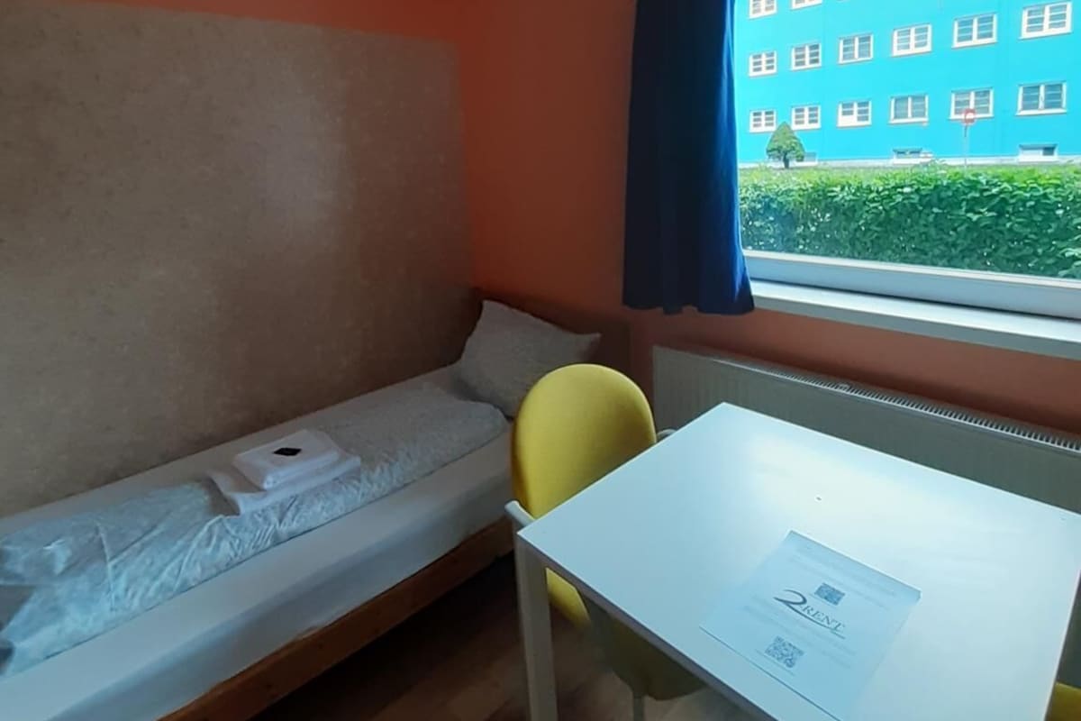 Une petite pièce avec un lit simple, une table et une chaise. Il y a une fenêtre donnant sur un immeuble et sur la verdure environnante.
