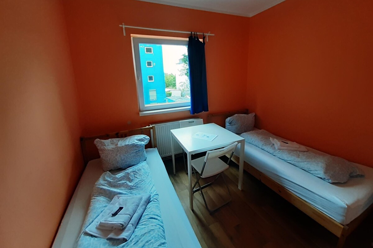 Une petite chambre avec deux lits simples, une petite table et une fenêtre donnant sur un immeuble bleu.