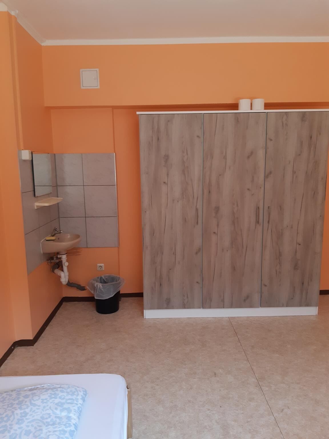A bedroom with a bed, a wardrobe, a sink, and a trash bin.