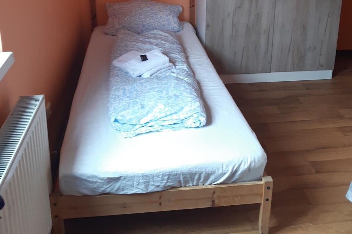 Une petite chambre avec un lit simple, une tête de lit en bois, un matelas blanc et une couverture grise à motifs. Une grande armoire en bois clair est adossée au mur.