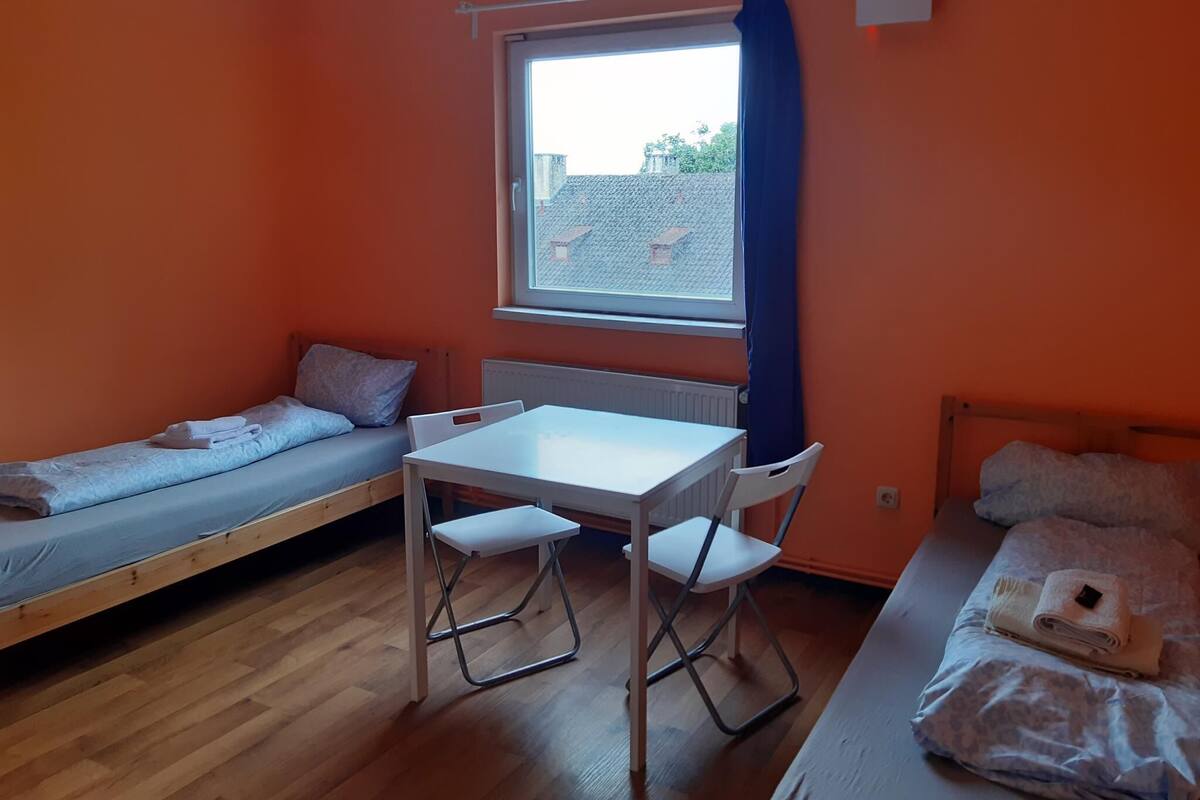 Una habitación pequeña con dos camas individuales, una mesa y sillas. Se ve una ventana con vistas a edificios.