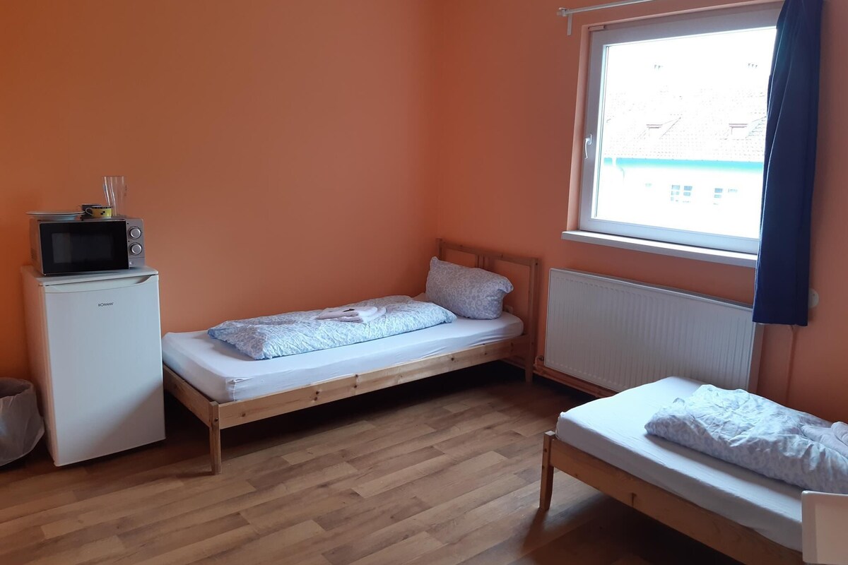 Une petite pièce aux murs orange, avec deux lits simples, un petit réfrigérateur blanc et une fenêtre donnant sur des immeubles.