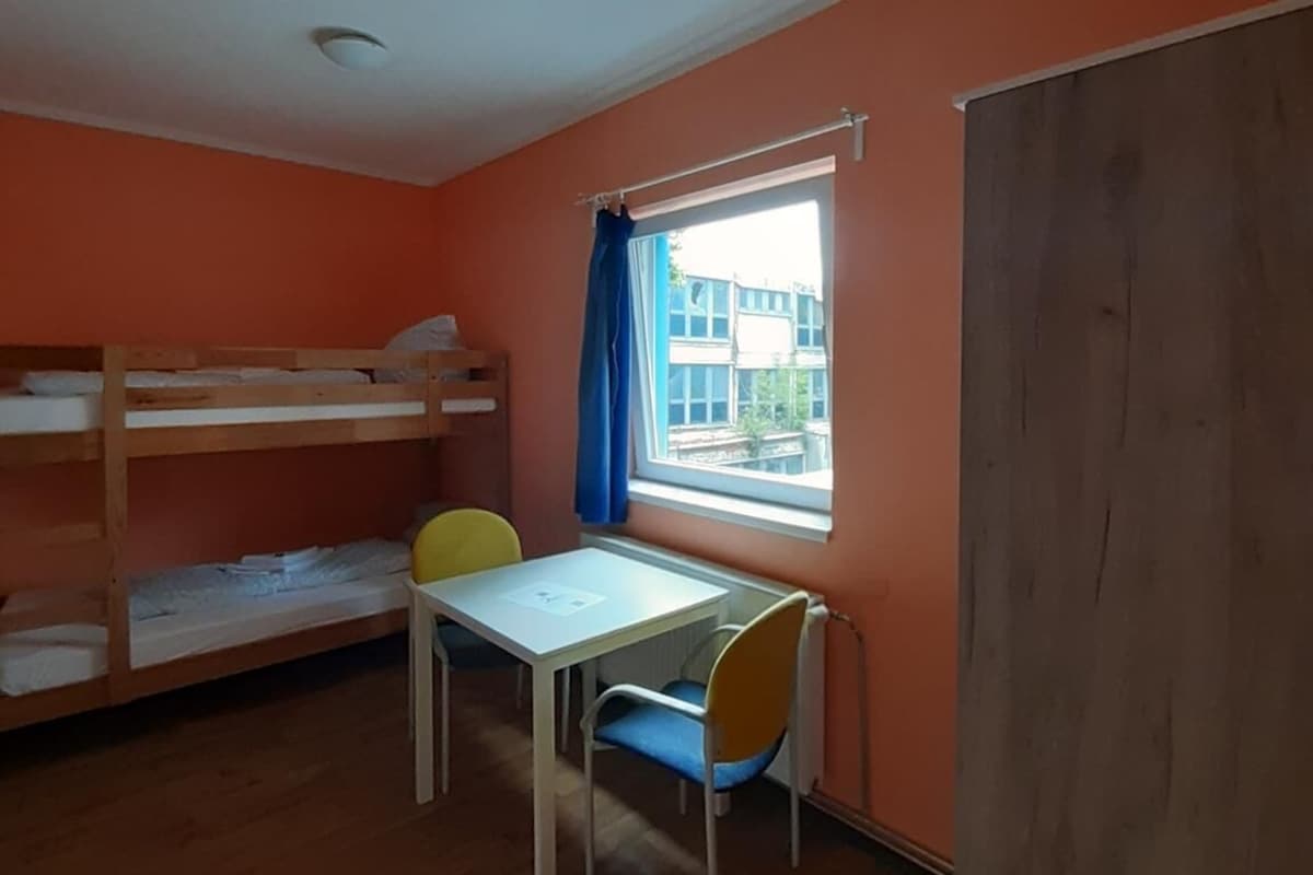 Une chambre avec un lit superposé, une table avec des chaises, une fenêtre avec des rideaux et un radiateur.