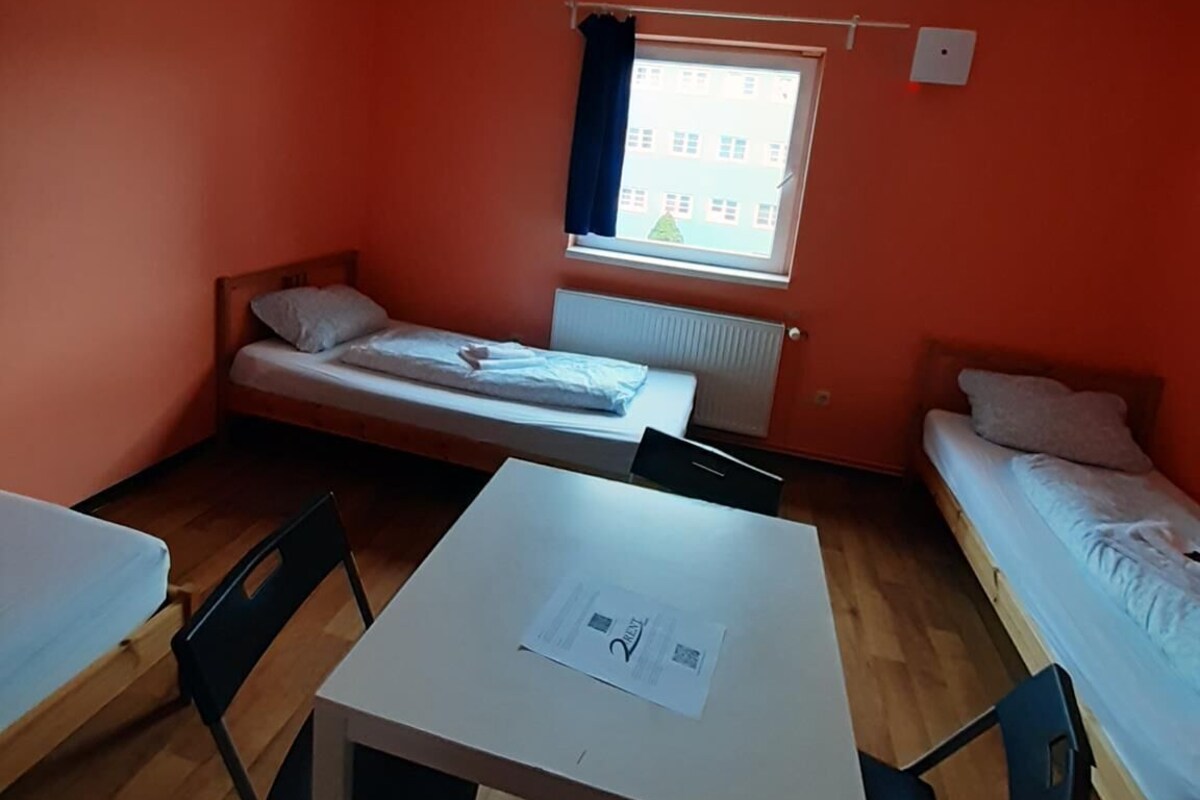 Ein Schlafsaalzimmer mit zwei Einzelbetten, einem kleinen Tisch mit Stühlen und einem Fenster mit Vorhängen.