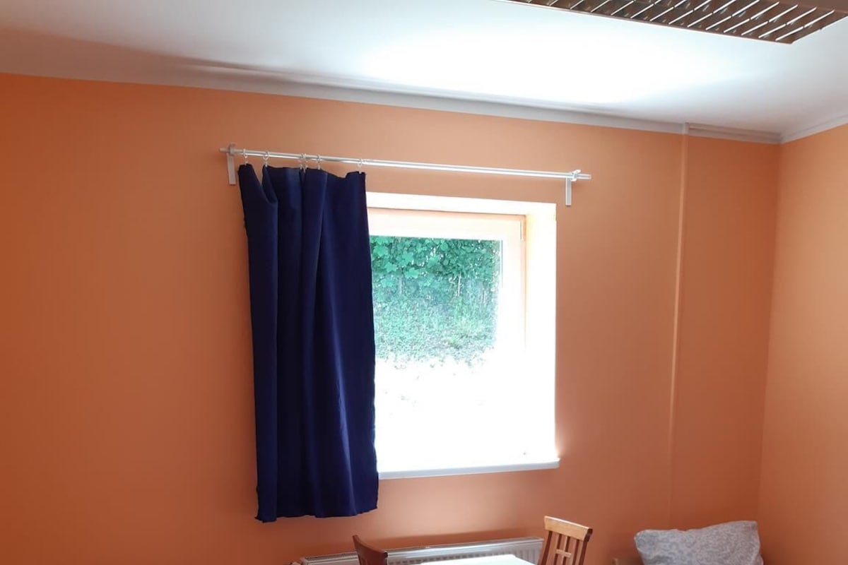 Une petite pièce lumineuse avec un mur orange, une fenêtre ornée d’un rideau bleu, un lit simple, une table avec des chaises et un radiateur.