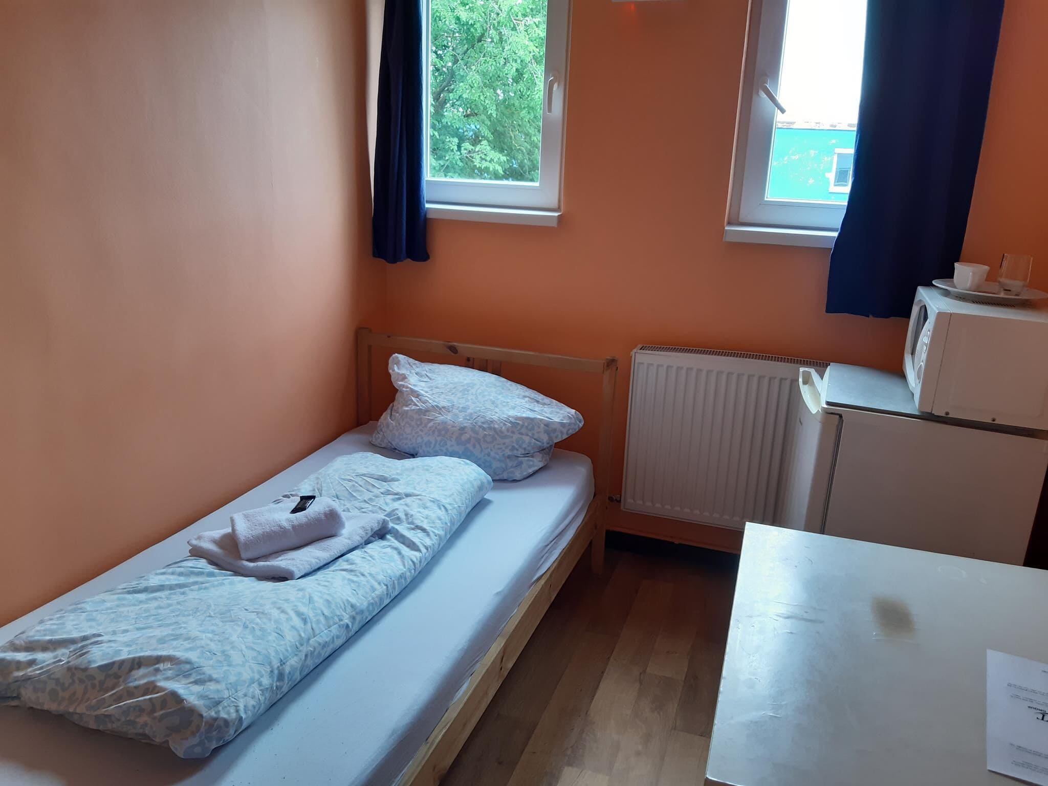 A small, single-bed room with a wooden frame, a white mattress, a white pillow, a white blanket, a white radiator, and a white table with a cup and saucer.