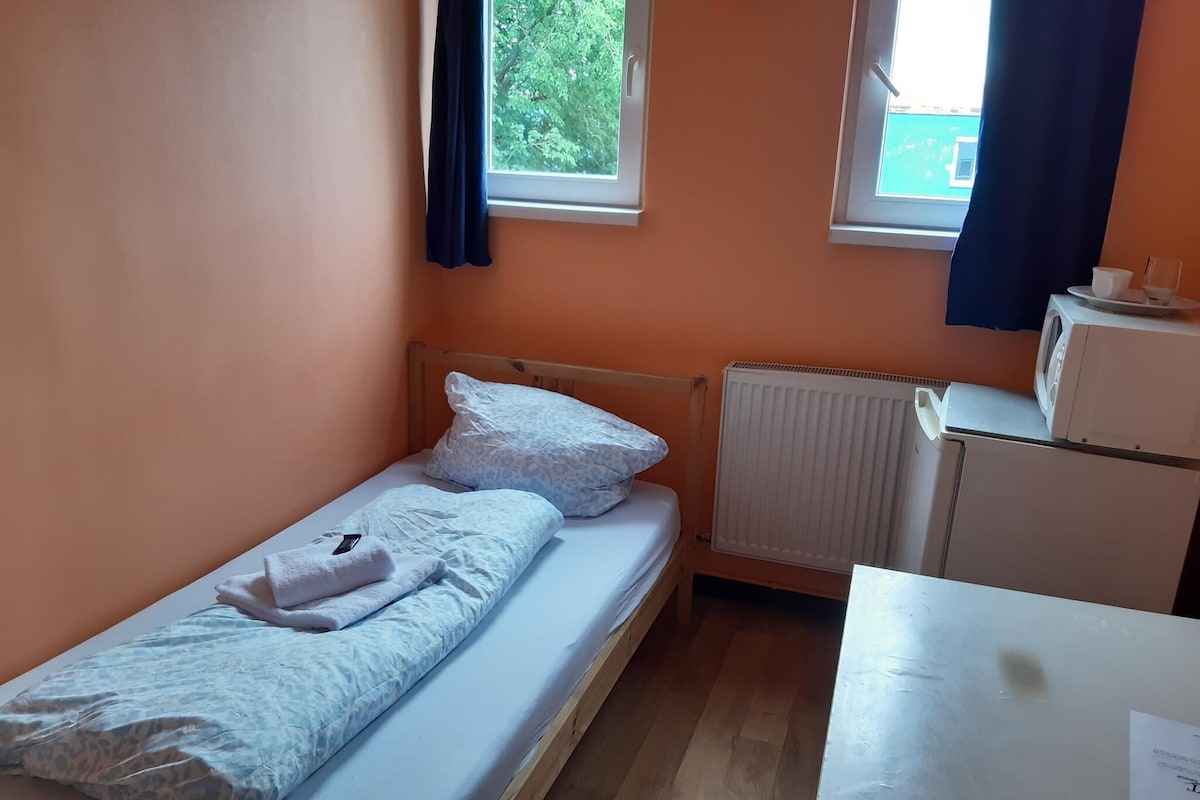 Une petite chambre avec un lit simple, un cadre en bois, un matelas blanc, un oreiller blanc, une couverture blanche, un radiateur blanc et une petite table blanche sur laquelle se trouvent une tasse et une soucoupe.