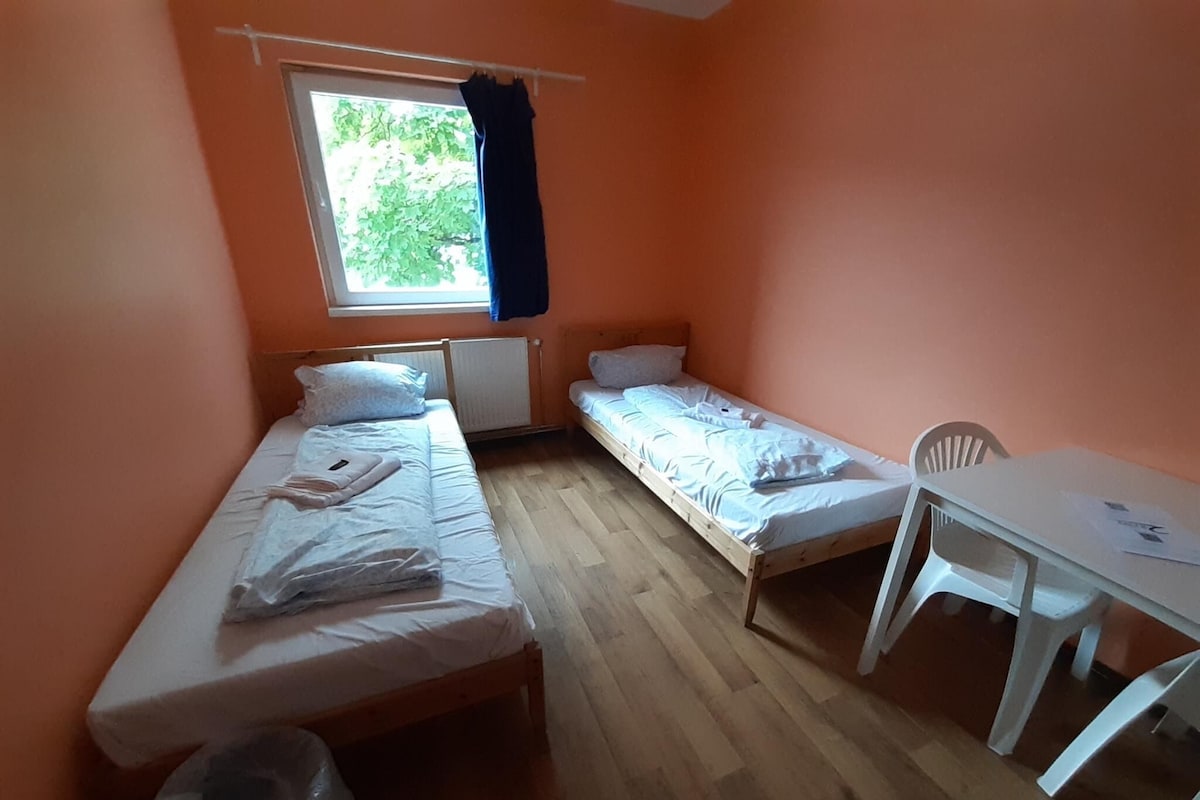 Une chambre de dortoir avec deux lits simples, une petite table et une fenêtre avec des rideaux.