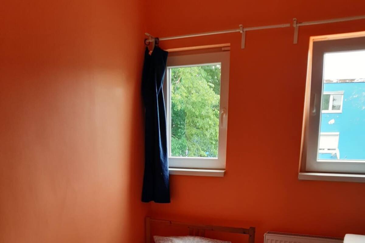 Een kleine eenpersoonskamer met een oranje muur, een raam met uitzicht op groen, een radiator en een houten tafel.