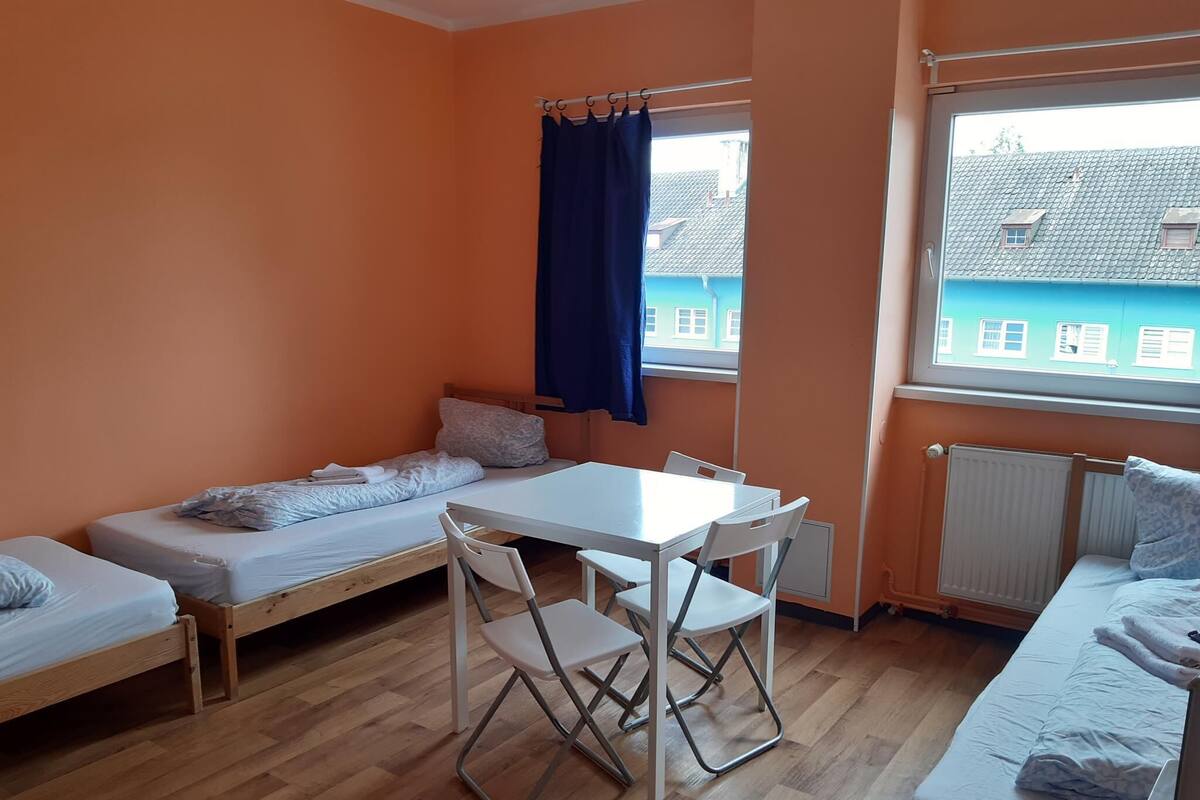 Una habitación pequeña y luminosa con dos camas individuales, una mesa con sillas y un ventanal con vistas a edificios.