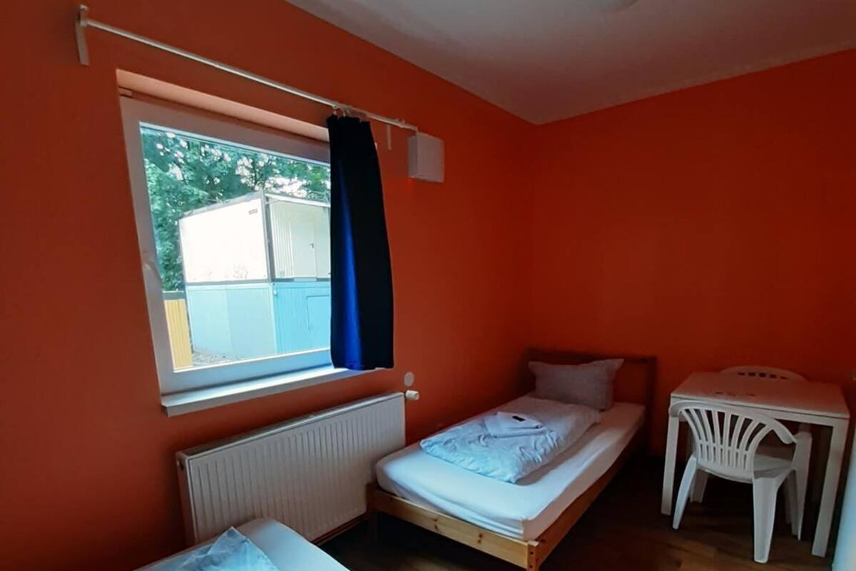 Een kleine, lichte kamer met twee houten bedden, een witte tafel en een raam met uitzicht op een gebouw.