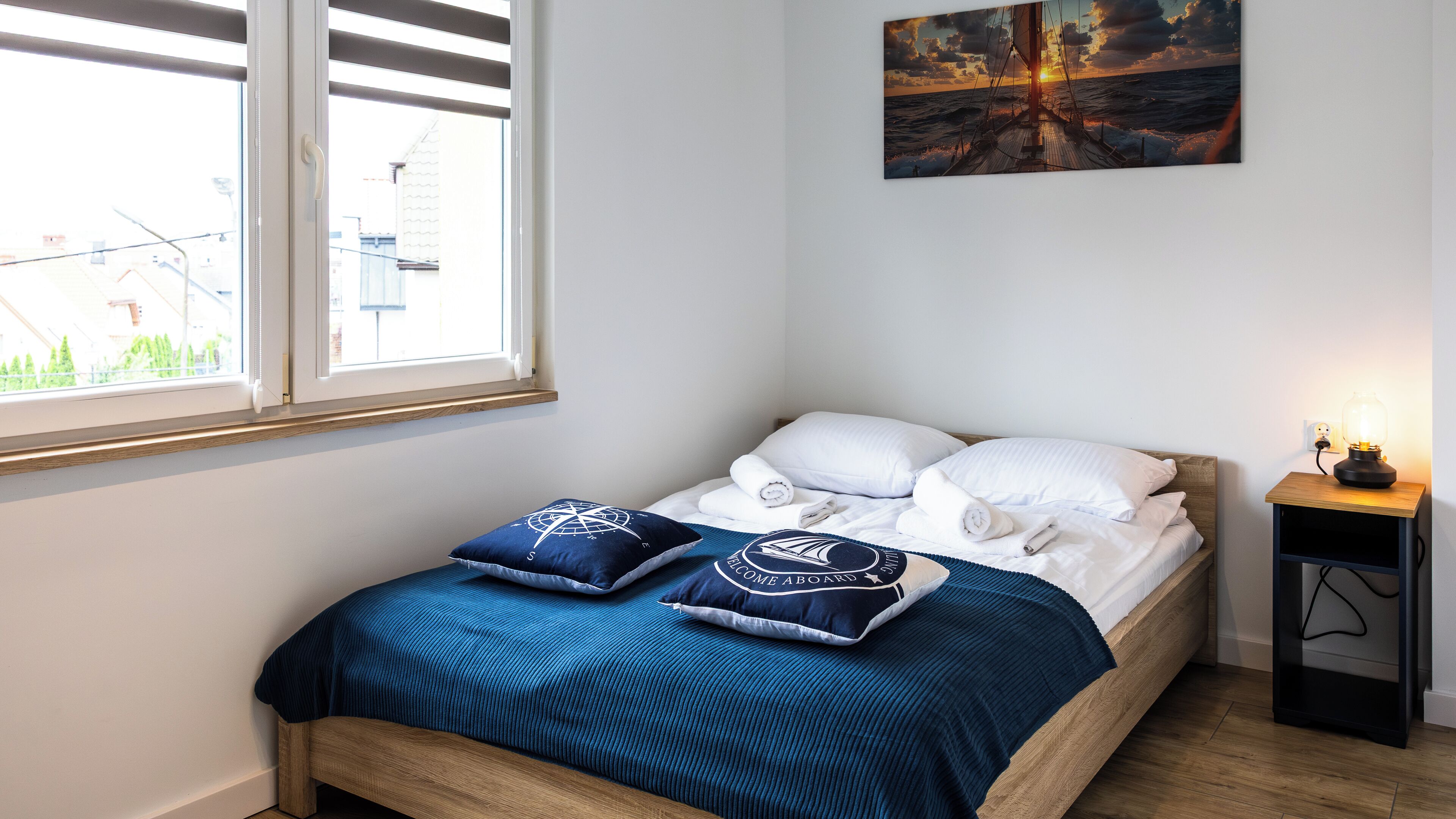 Un lit bien fait, avec une couvre-lit bleue et des coussins assortis, une petite table de chevet avec une lampe, et un tableau représentant un coucher de soleil sur l’océan accroché au mur.