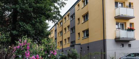 Un edificio de varios pisos con balcones, un jardín con flores rosadas y un coche estacionado.
