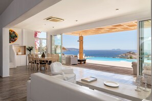 A modern interior with a dining area, white furniture, and a view of the ocean. - Alegria Mykonos 10 Suites – Luxury Estate with 14 Suites and Panoramic Sea Views (Kalafatis)