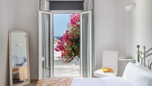 A hotel room with a bed, a chair, a bedside table, a mirror, and a view of the sea through an open door. - Alegria Mykonos 10 Suites – Luxury Estate with 14 Suites and Panoramic Sea Views (Kalafatis)