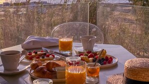 Una mesa dispuesta con alimentos para el desayuno, una bebida y un sombrero de paja, con vistas a la costa.