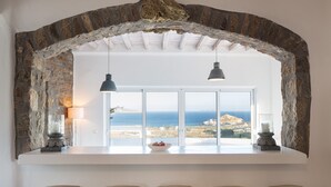 Una ventana enmarca con piedra que da al mar, una barra blanca con taburetes y lámparas colgantes.