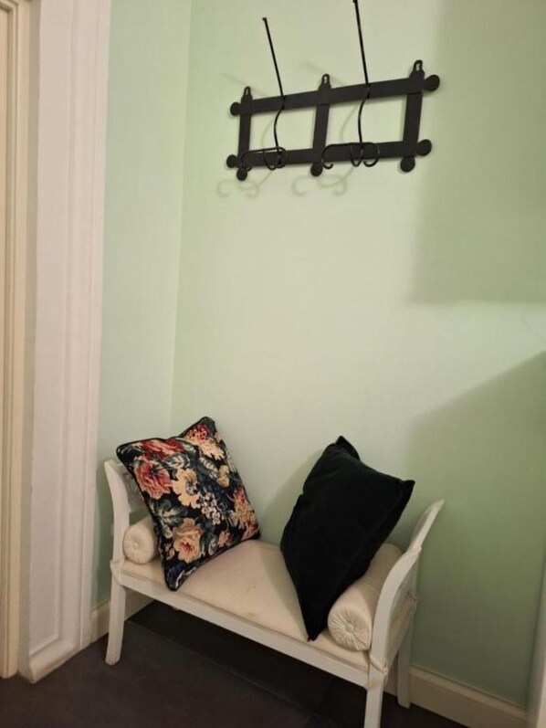 Un banc blanc, agrémenté d’un coussin à motifs floraux et d’un coussin noir, est adossé à un mur vert clair. Au-dessus, on aperçoit une étagère en métal noir.