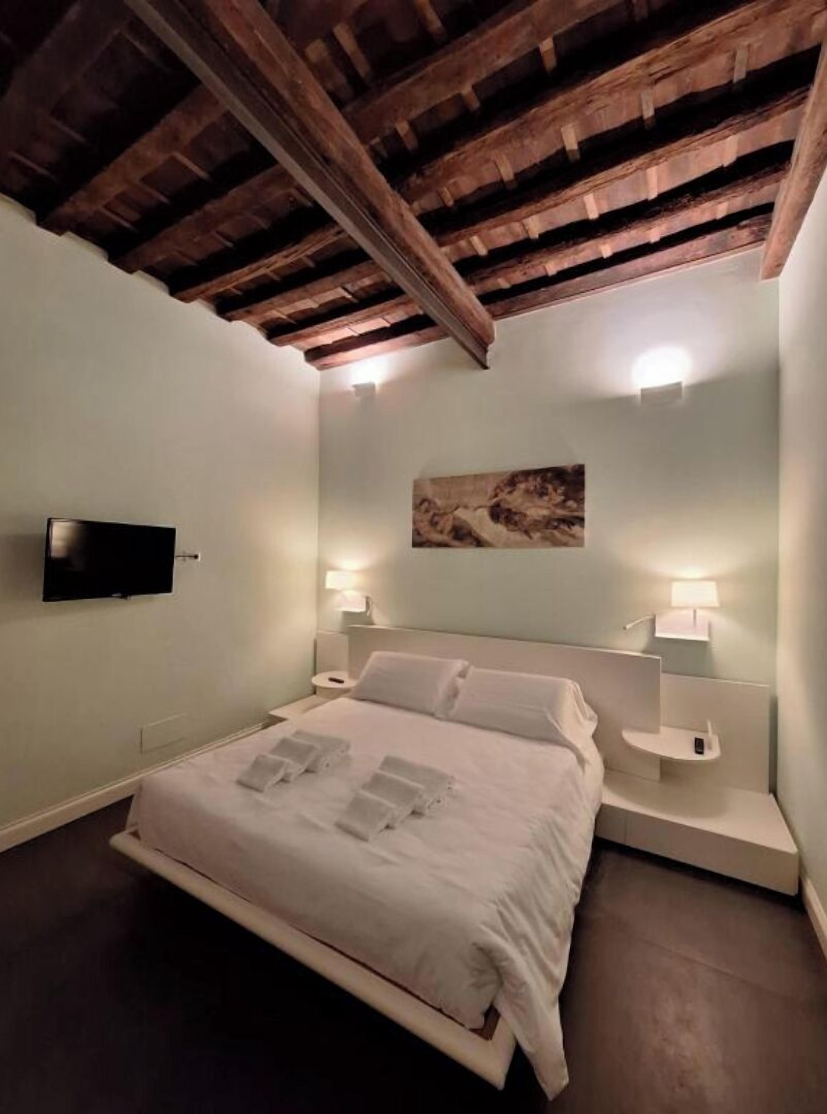 A bedroom with a wooden ceiling, a bed with white linens, two bedside tables with lamps, and a mounted TV.