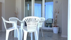 Ein Balkon mit weißen Plastikstühlen und einem Tisch, einem Blumentopf und einer Glastür mit Vorhängen.